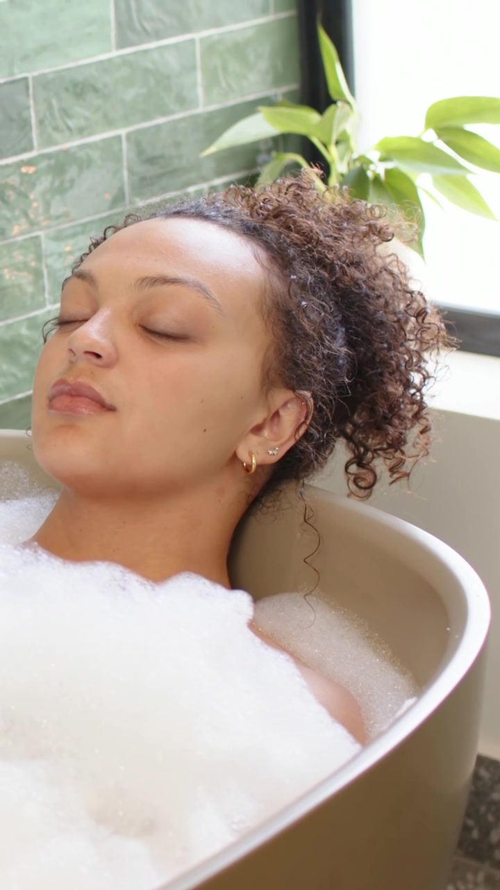 Vertical video: Soaking to relax, woman breathing and shifting in freestanding tub at home, foam