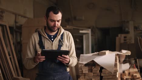 Un-Joven-Con-Barba-Camina-Por-El-Taller-De-Carpintería-Con-Una-Tableta-En-Las-Manos