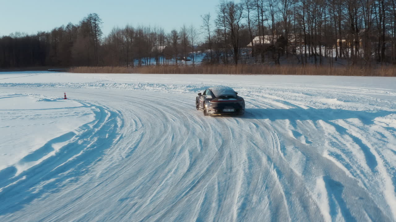 Drone footage of blue Porsche carrera 2020 911 4S slowly driving on frozen Lake in circle shaped track. Winter morning time, lot of snow particles, sunlight visible. Filmed in 4K resolution.