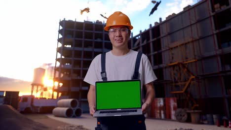 un trabajador asiático muestra una computadora portátil de pantalla verde a la cámara en el sitio de construcción