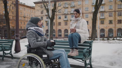 Mujer-Musulmana-Y-Su-Amiga-Discapacitada-En-Silla-De-Ruedas-Bebiendo-Café-Para-Llevar-En-Un-Banco-En-La-Ciudad-En-Invierno-6