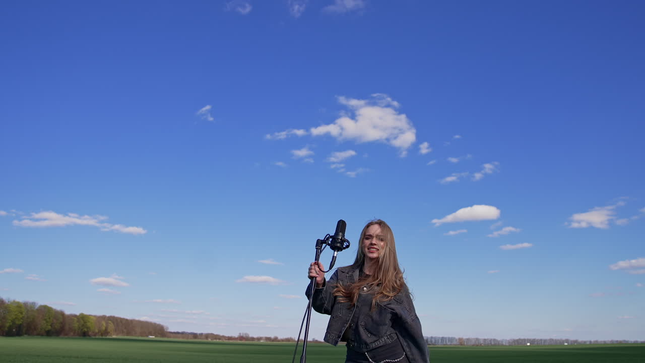 Beautiful young singer dancing with a microphone on a field. Pretty female soloist singing and jumping cheerfully outdoors. Slow motion.
