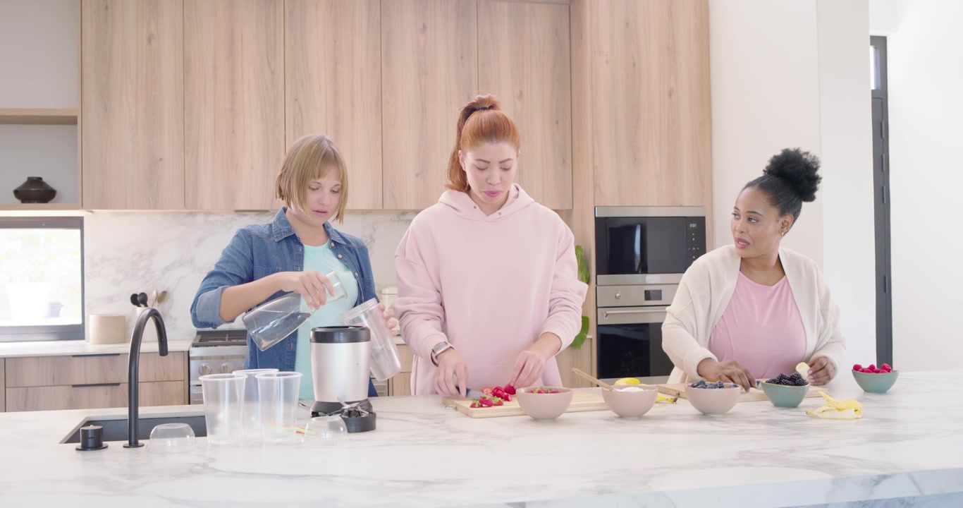 Diverse female friends prepping at island, blonde filling blender, others slicing fruit to blend