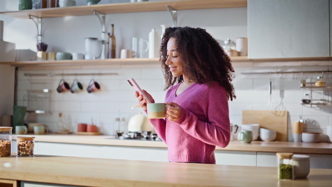 Casalinga felice che legge un messaggio sul cellulare in cucina, con un caffè in mano, primo piano.
