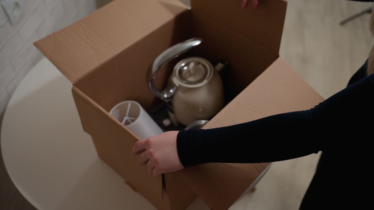 Unpacking kettle and mugs on table, box placed on round white table then opened to reveal kettle and cups, hands gently remove items, apartment interior, settling into new home, careful arrangement