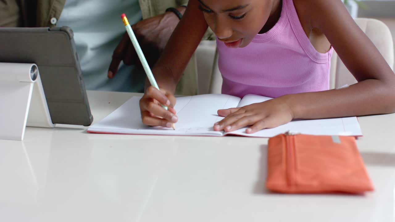 African American man supervising girl child writing in notebook at table, tablet guiding work