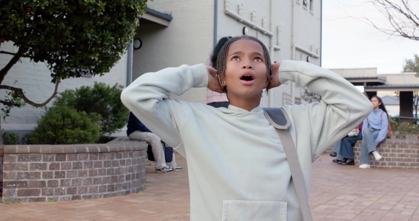 Teenage boy spotting above, looking up, raising arms, holding shoulder bag in school courtyard
