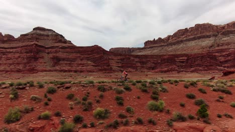 Toma-Aérea-De-Seguimiento-A-La-Izquierda-De-Un-Ciclista-De-Montaña-En-Bicicleta-A-Lo-Largo-De-Un-Sendero-Plano-En-El-Desierto-Rojo-De-Moab