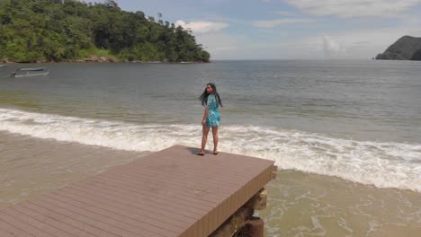 Aerial-drone-shoot-of-a-girl-walking-and-posing-on-a-jetty-with-fishing-boats-in-the-background