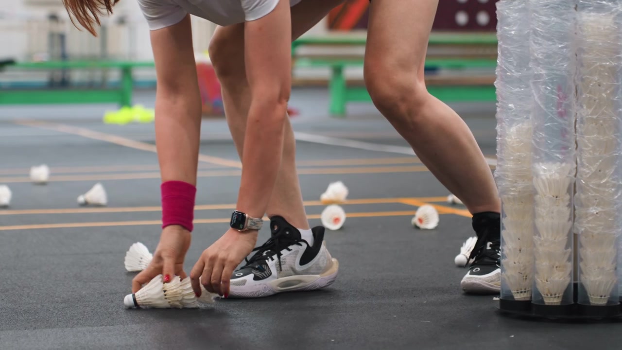 Jogador recolhendo petecas em quadra interna, mãos e pernas visíveis enquanto atleta pega petecas espalhadas do chão e de tubo plástico, munhequeira rosa, shorts pretos, tênis brancos, treinamento focado