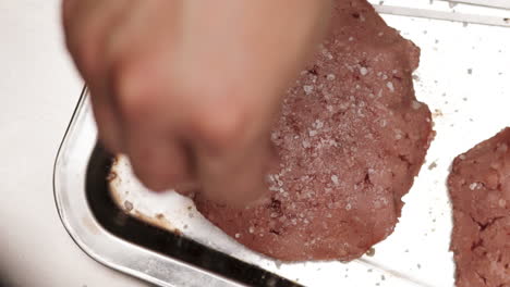 hand sprinkling rock salt on uncooked burger patties