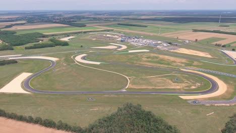 an aerial video of a modern racetrack complex winding through a wide open landscape of fields and forests. the track includes sharp curves, run-off zones, and support buildings with some bikes