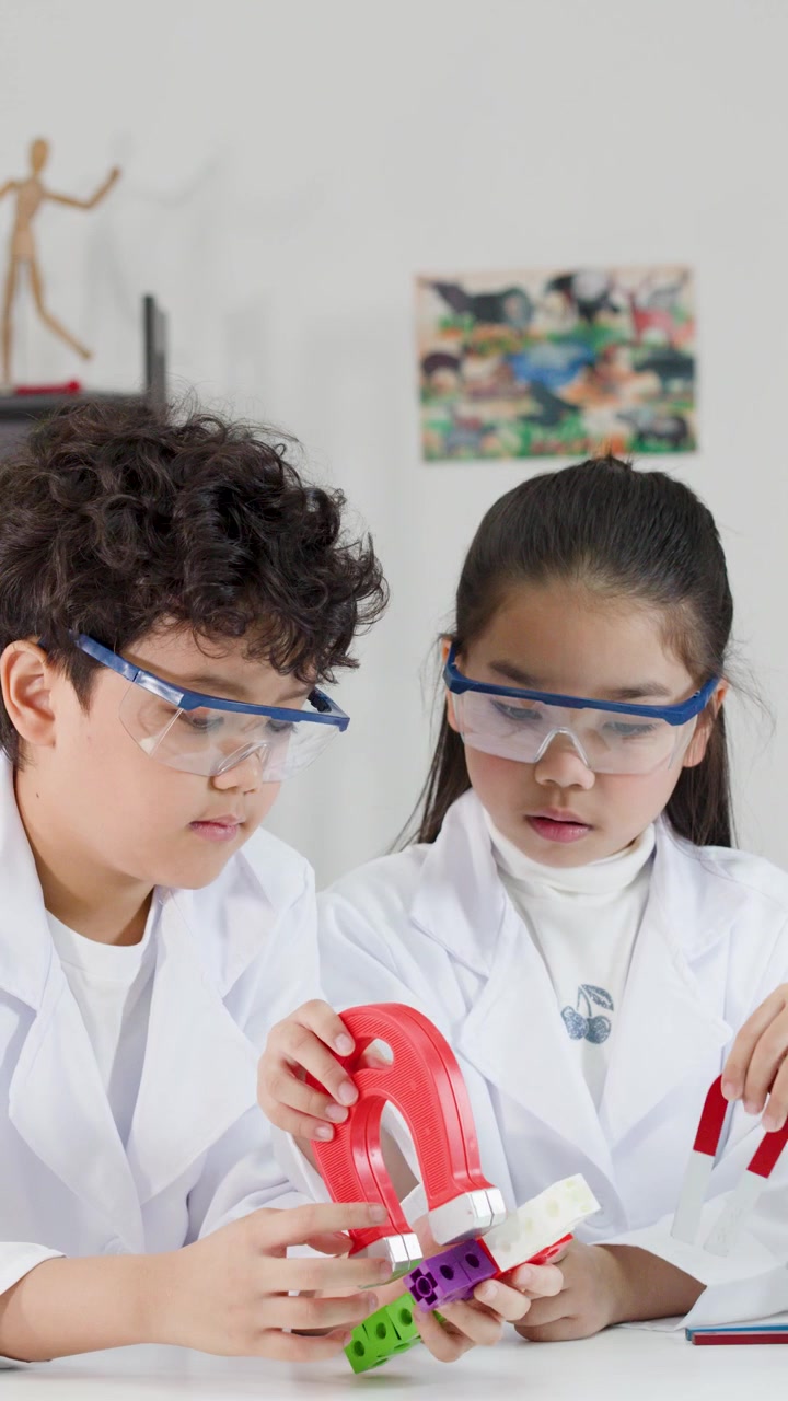 Dua siswa muda mengenakan kacamata pengaman menjelajahi magnetisme menggunakan magnet merah besar di dalam kelas