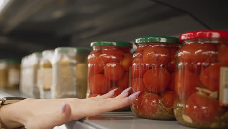 close up of demure shopper hands touching glass jar of tomatoes on metal shelf rails in bright grocery aisle highlighting manicure polished nails and product selection with blurred retail background