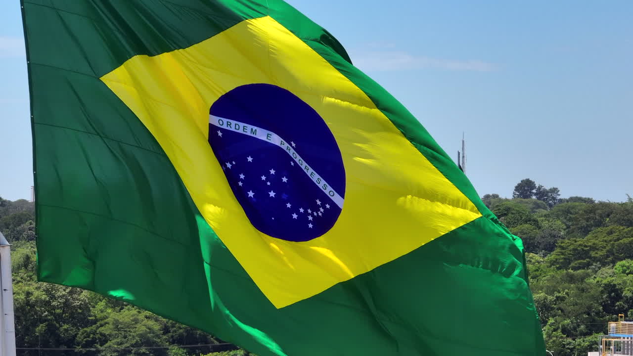 Duas bandeiras nacionais do Brasil tremulando ao vento com uma bandeira grande em primeiro plano e uma segunda bandeira ao fundo contra um céu azul e paisagem verde.