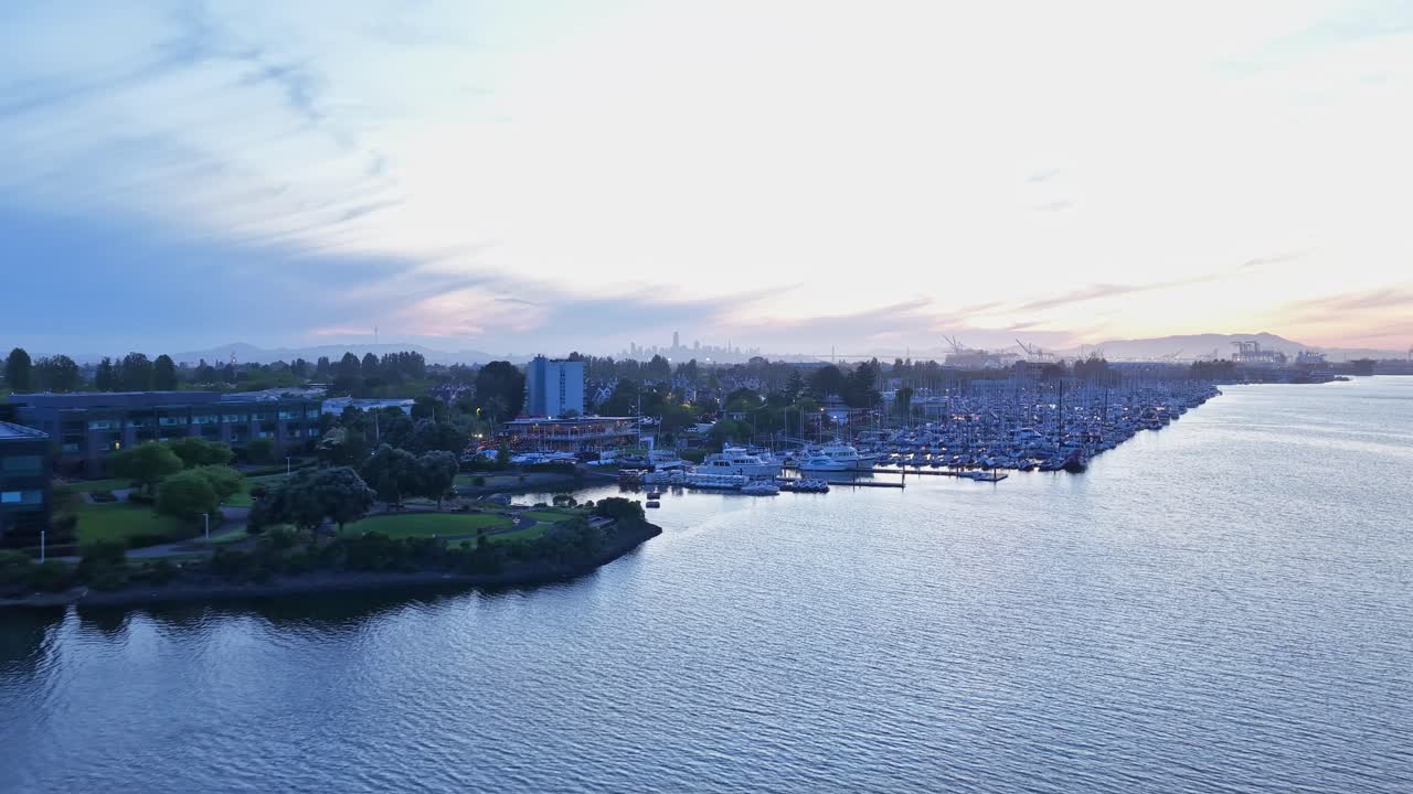 Luftfoto med zoom inn over Encinal-bassenget og marinaen i Alameda, California, under en fredelig solnedgang, med San Francisco- og Oakland-skyline synlig i den fjerne horisonten
