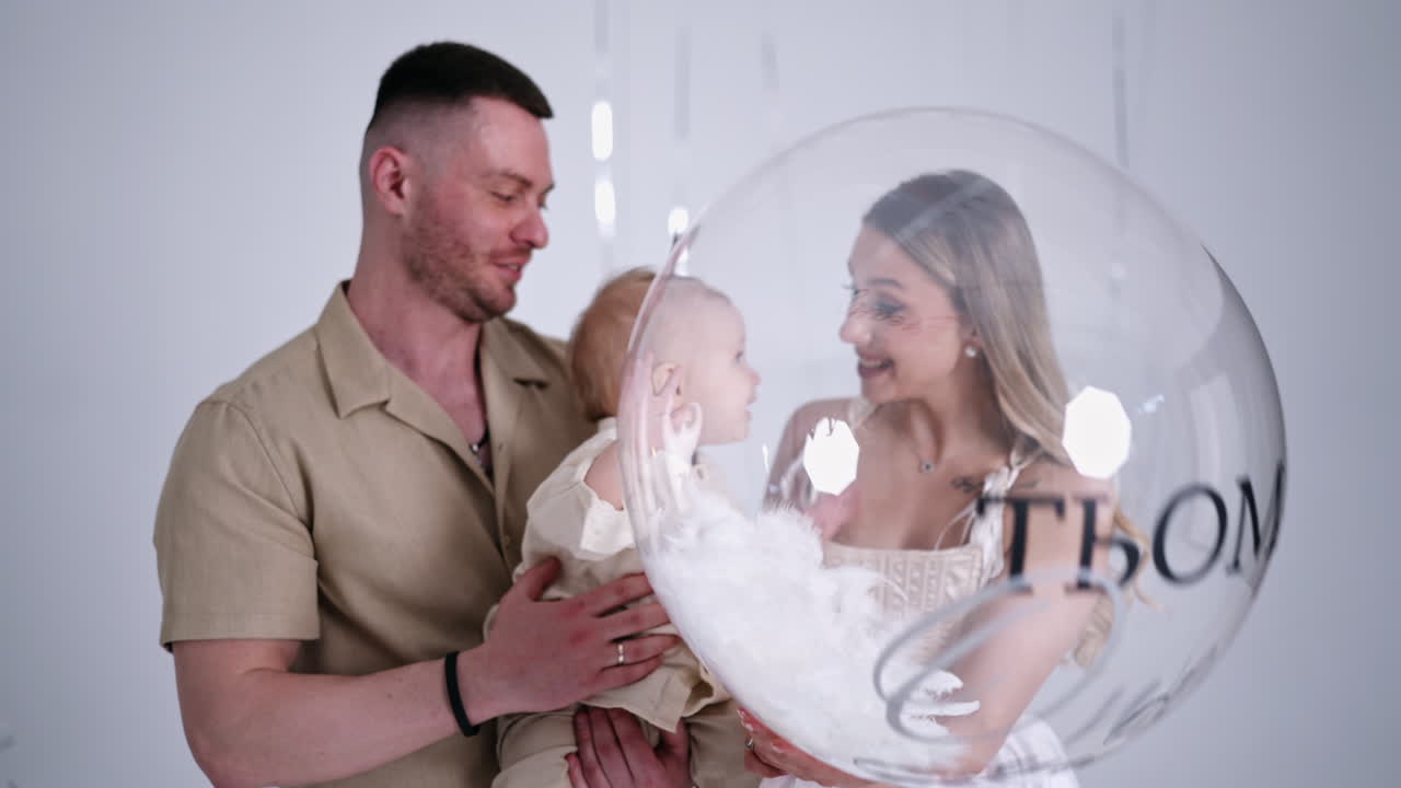 Happy one-year-old toddler holding a big transparent balloon. Portrait of a beautiful Caucasian family in studio.