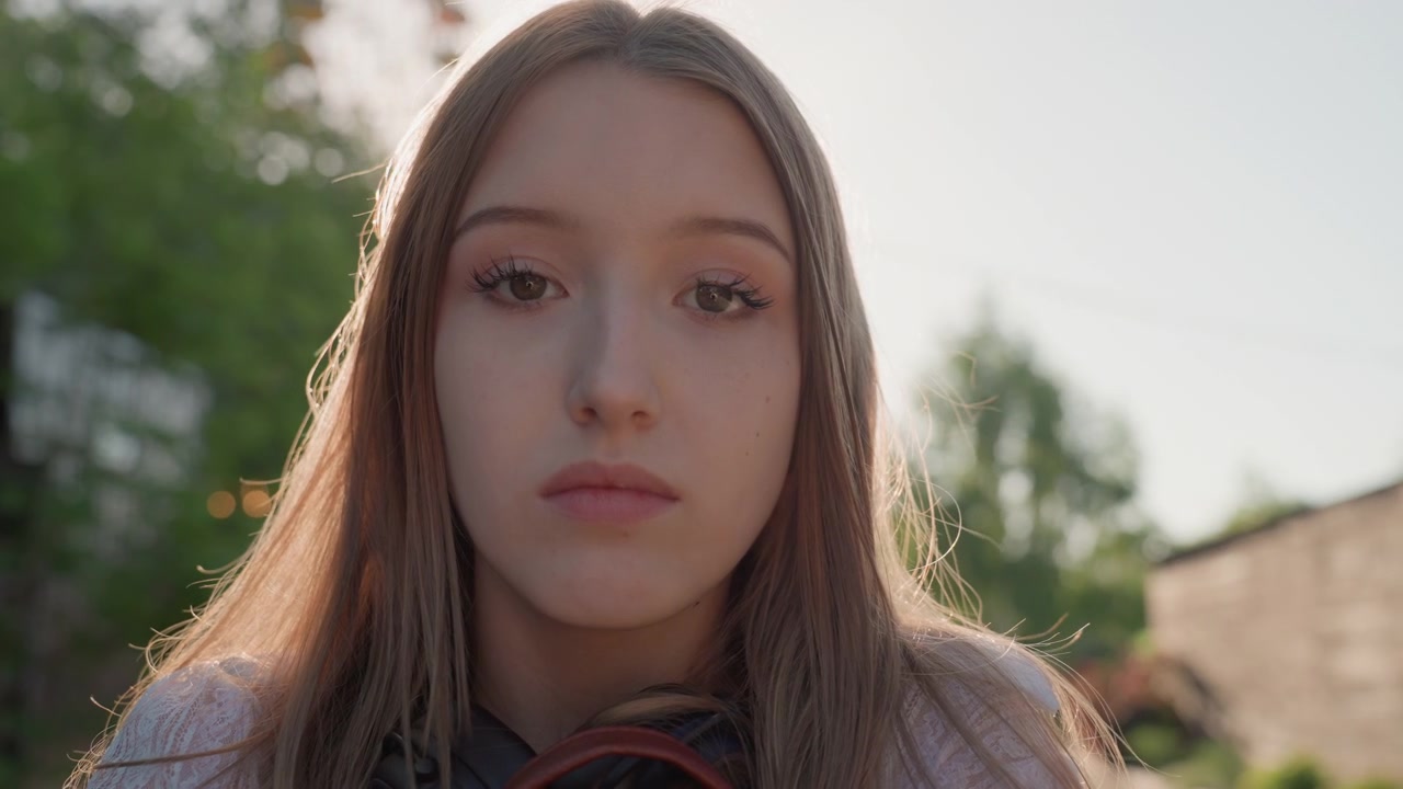 Primo piano di una giovane donna con lunghi capelli in piedi all'aperto sotto una luce solare soffusa, che indossa abiti casual. La sua espressione è calma e contemplativa mentre guarda in lontananza, suggerendo tranquillità