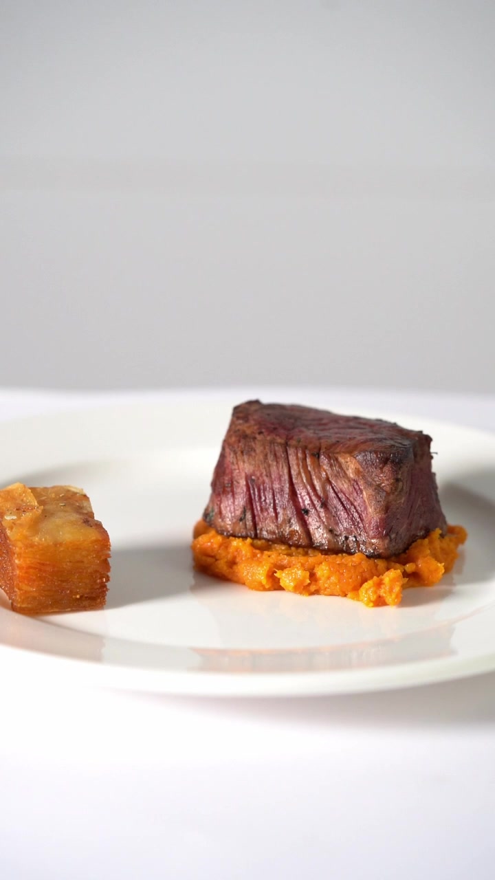 Succulenta bistecca di manzo alla griglia servita su purè di verdure cremoso in un piatto minimalista, primo piano.
