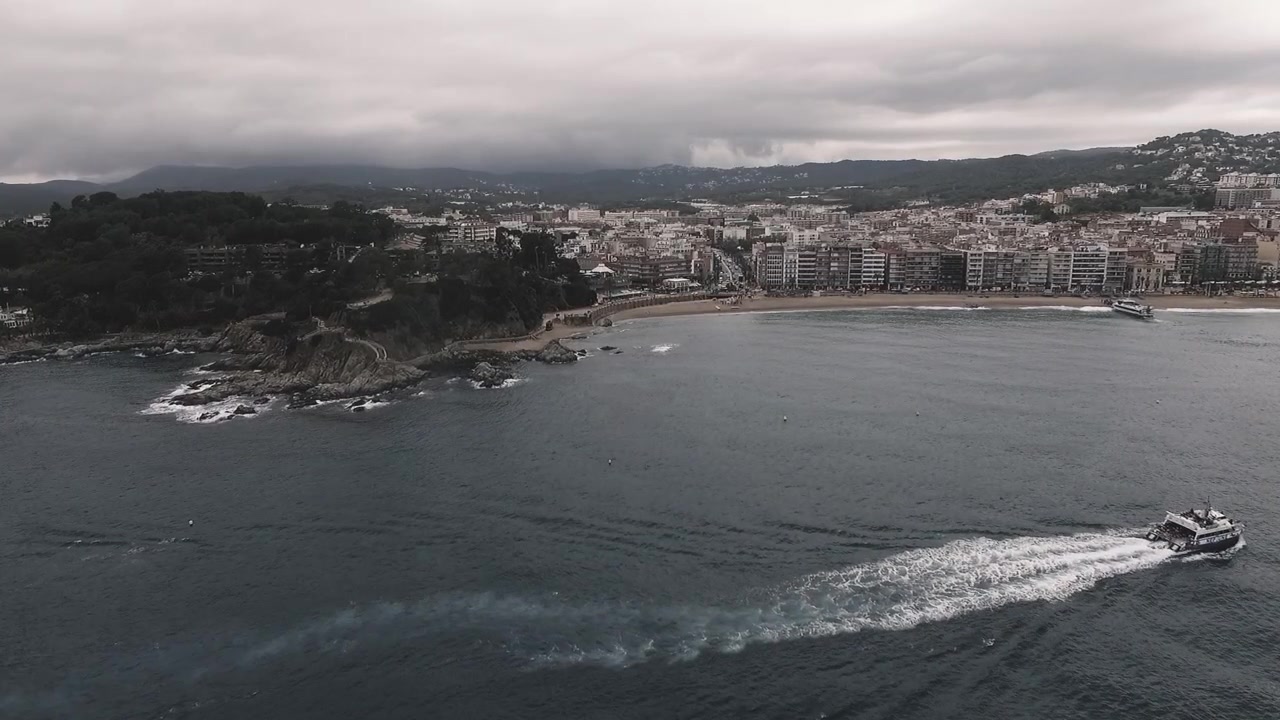 Ripresa aerea con drone di un motoscafo che naviga lungo la costa della Costa Brava con una città costiera mediterranea e nuvole spettacolari sullo sfondo.