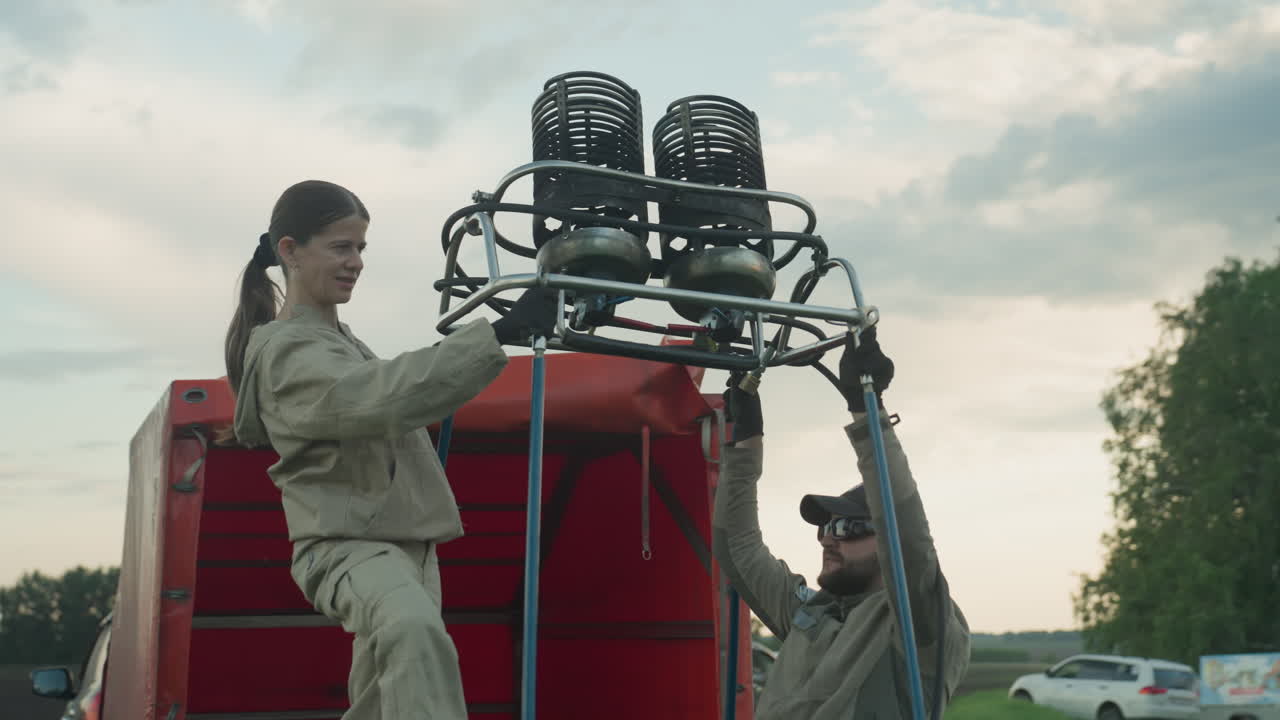 close up of couple aligning twin burner assembly into wicker balloon basket beside red trailer on grassy plain under cloudy sky, focus on precise teamwork and safety setup before flight prep