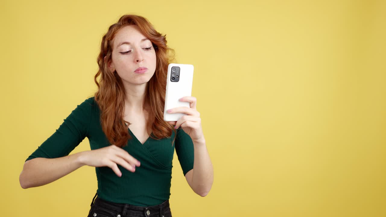 Young woman checking her appearance or taking a selfie with a smartphone against a yellow background