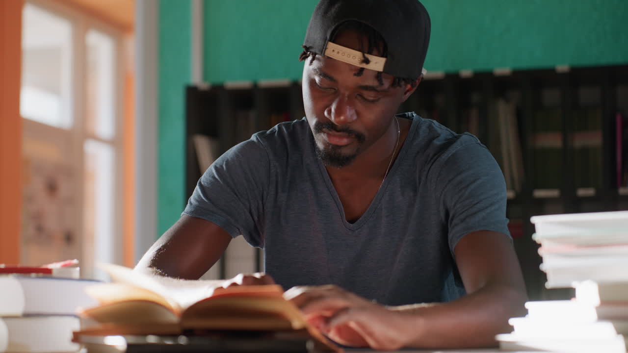 Young man in library stretches arms after intense studying, eyes closed in relief beside open book and stacks, taking short break at desk as warm sunlight filters through window