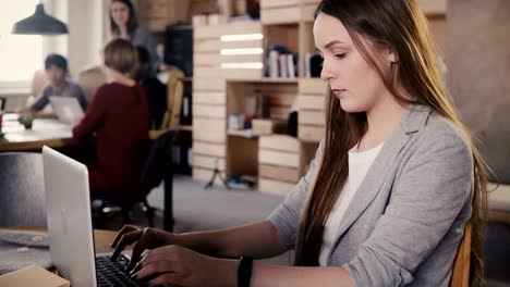 concentrated european young girl typing a letter on laptop. hard work in trendy office space, teamwork in background 4k