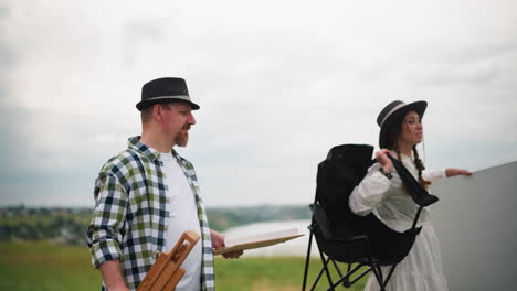 a man and woman walk and talk in a grassy field. the man, wearing a hat and holding a white cardboard box, converses with the woman in a white dress and hat