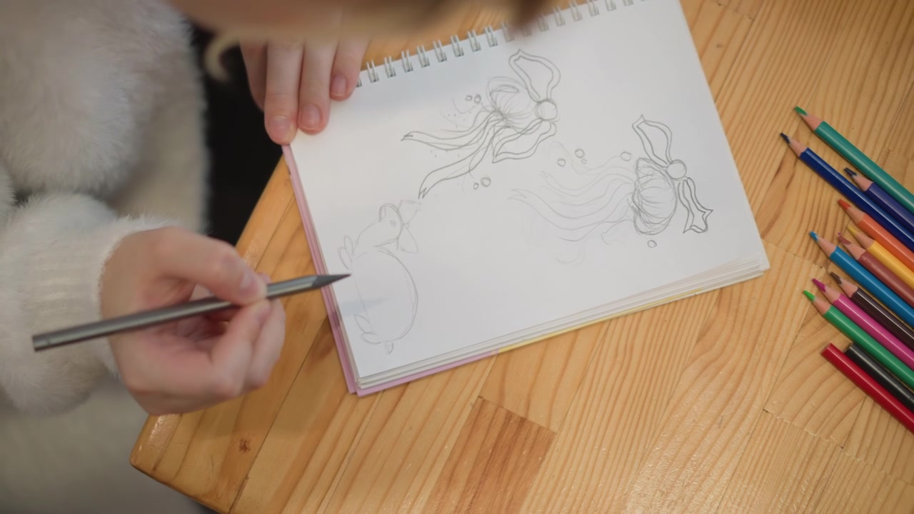 Primo piano dall'alto di un bambino che disegna su un quaderno a spirale aperto su un tavolo di legno, la mano destra guida la penna sulla pagina mentre la sinistra tiene fermo il foglio, con scarabocchi e matite colorate nelle vicinanze.