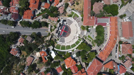 drone top down view of plovdiv's ancient theatre of philippopolis, showcasing its circular architecture amidst urban surroundings. the vibrant red rooftops and lush greenery enhance the scenic vista