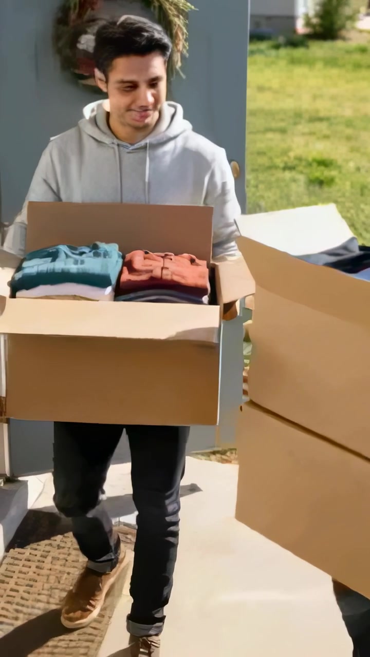 Vertical video: Stepping onto porch, man in gray hoodie carrying box moving inside to bring clothes