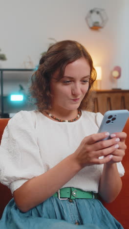 happy young woman sitting on sofa using smartphone texting smiling at home living room apartment