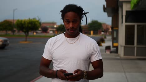 african american man with dreadlocks and in a white t-shirt walking and texting typing on his phone, and looking around, and into the camera laughing