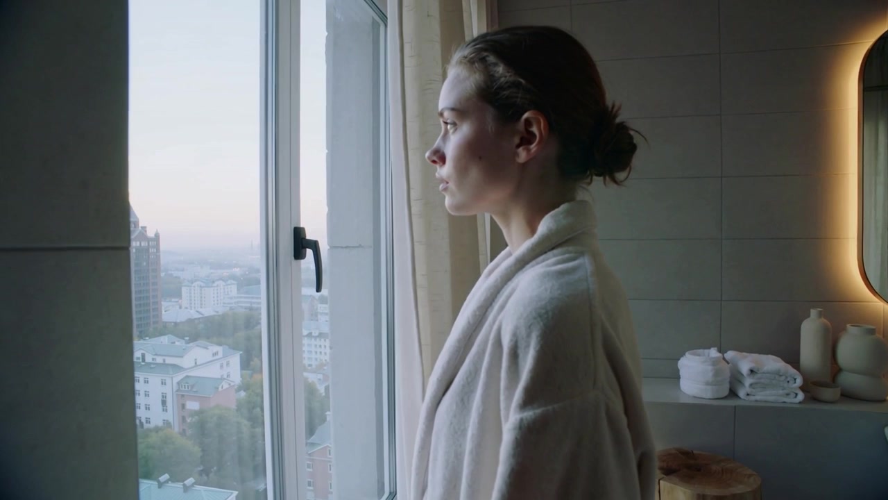 Tirando la tenda, una donna in accappatoio si avvicina alla finestra e osserva lo skyline in attesa della luce del mattino.
