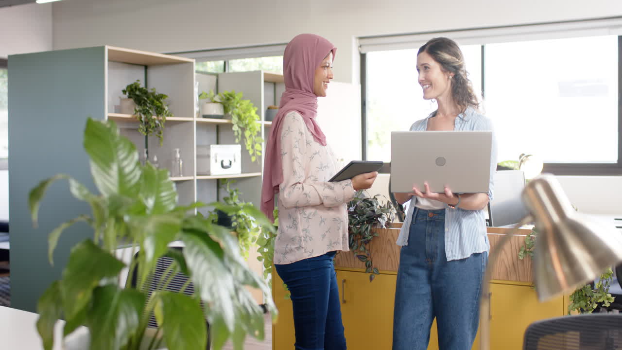 Diverse creative female colleagues in discussion using laptop and tablet in office, slow motion