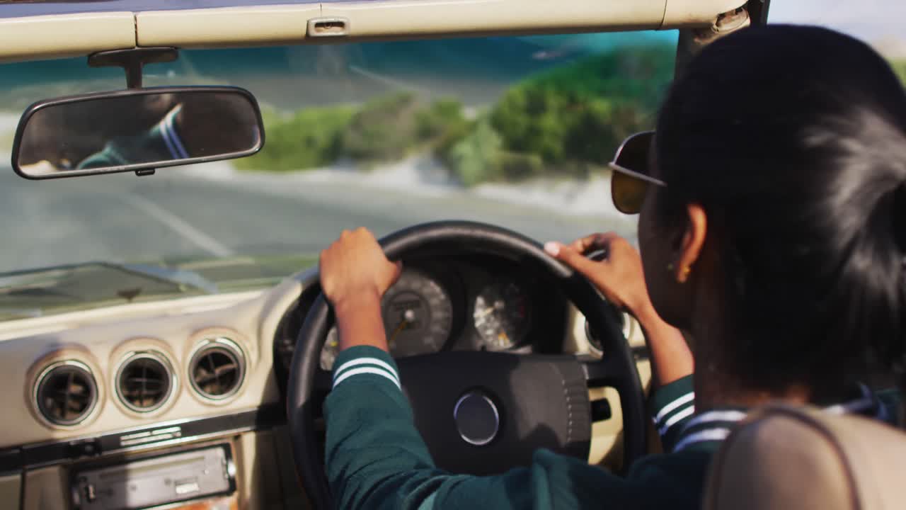 아프리카계 미국인 여성이 컨버터블 ca에서 시골 도로를 따라 운전하는 동안 뒤쪽 시야 거울을 조정합니다.