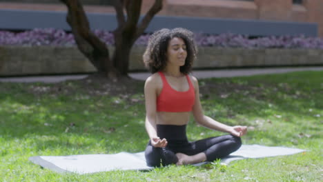 shot of a girl with curly hair doing meditation on the grass in the park while enjoying the clean air in the morning