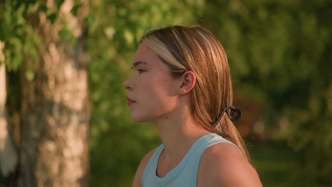 Close-up side view of young lady looking unhappy while moving gracefully outdoors like roller skating, with sunlight reflecting off her face and hair tied back, background includes lush natural trees