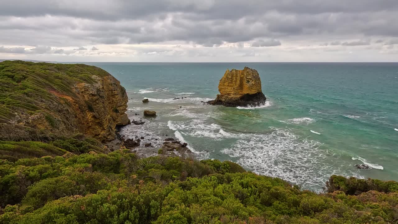 Wide shot of rugged coastline with prominent sea stack, green cliffs, and turquoise waves under overcast skies. Subtle camera pan reveals dramatic seascape