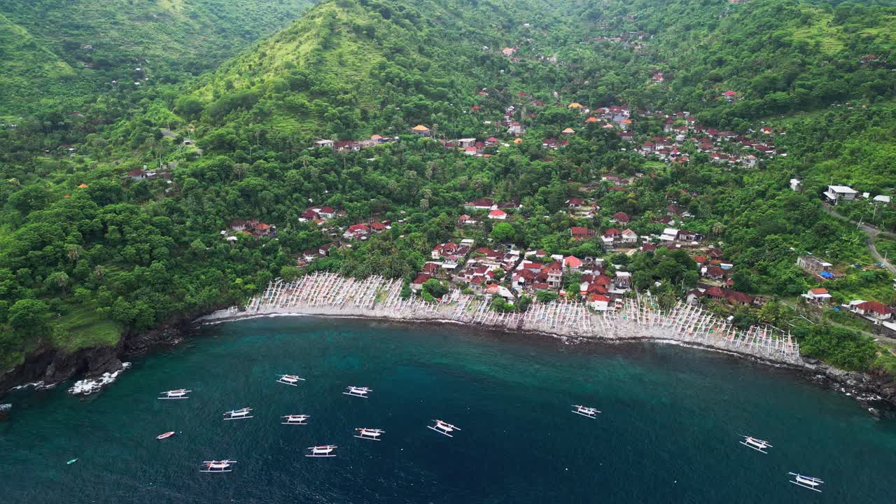 Un video aereo con drone cattura un denso villaggio tropicale costruito tra verdi colline e una costa curva, dove centinaia di tradizionali barche da pesca riposano su una spiaggia rocciosa accanto all'acqua blu profonda dell'oceano.