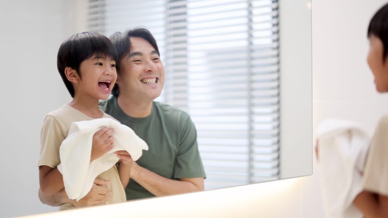 Father and son laughing together while brushing teeth in a bright, modern bathroom at home