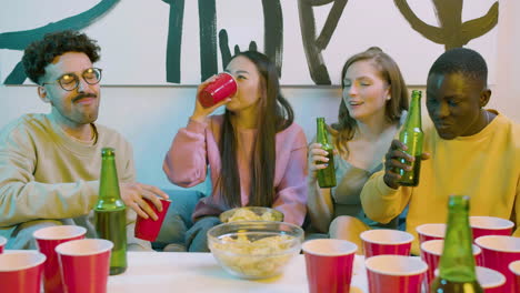 happy multiethnic young friends toasting with beer and eating potato chips while sitting on sofa at home