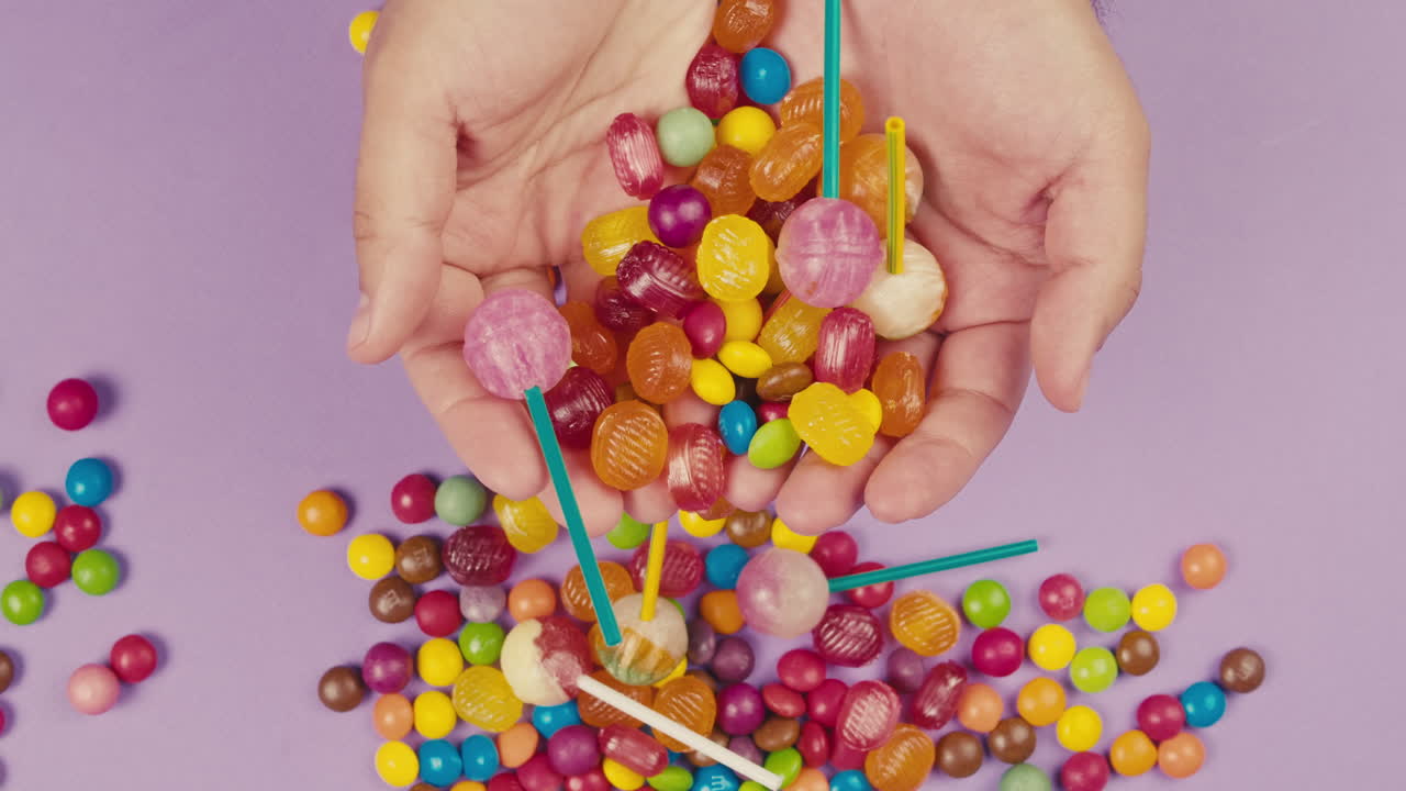 Colorful dragee candies, lollipops, bonbons in woman hands, purple background. Summer bright texture studio shot for store, supermarket, close-up top view. Mix of children sweets made of sugar.