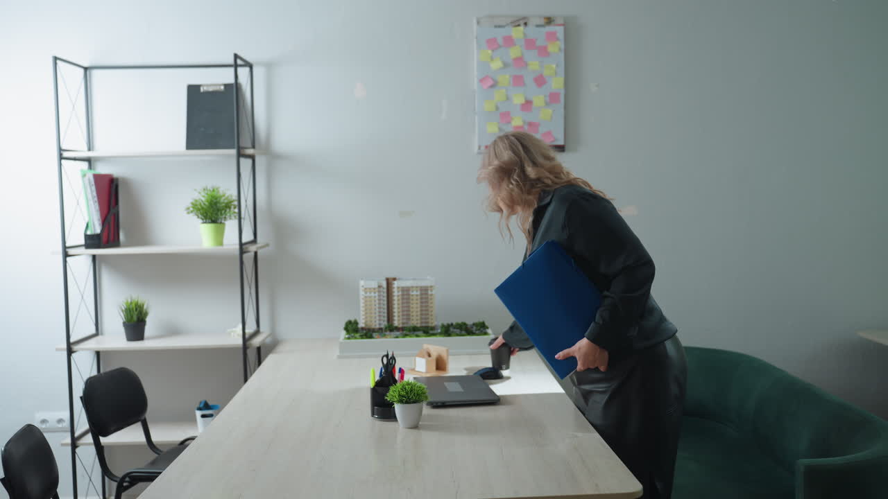Real estate expert enters office holding coffee cup and document folder, approaches desk to start day, setting items down on clean organized workspace with architectural model