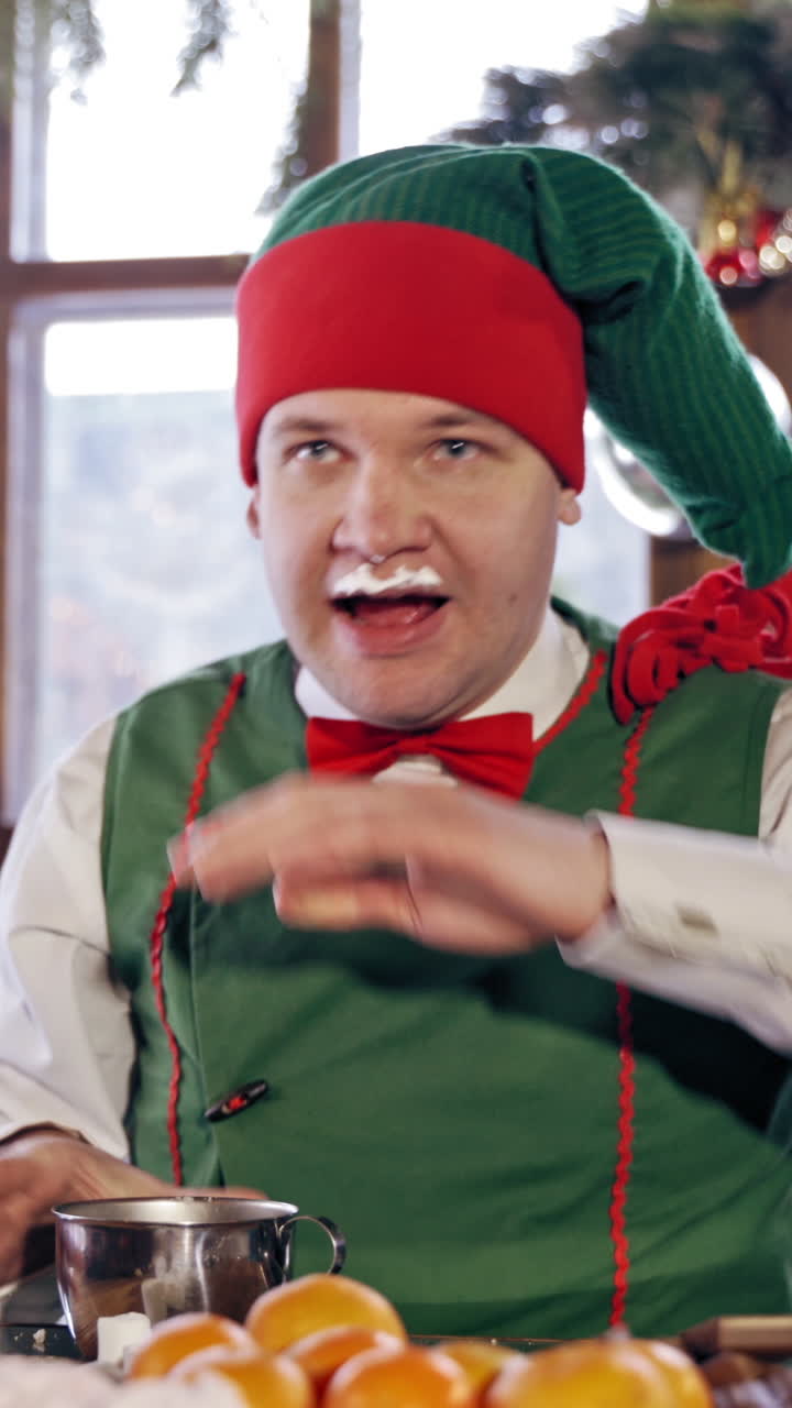 Funny elf with drinks and becomes dirty on his face on Christmas background. Hungry male elf in red and green costume drinks at festive table indoors. Close-up. Vertical video