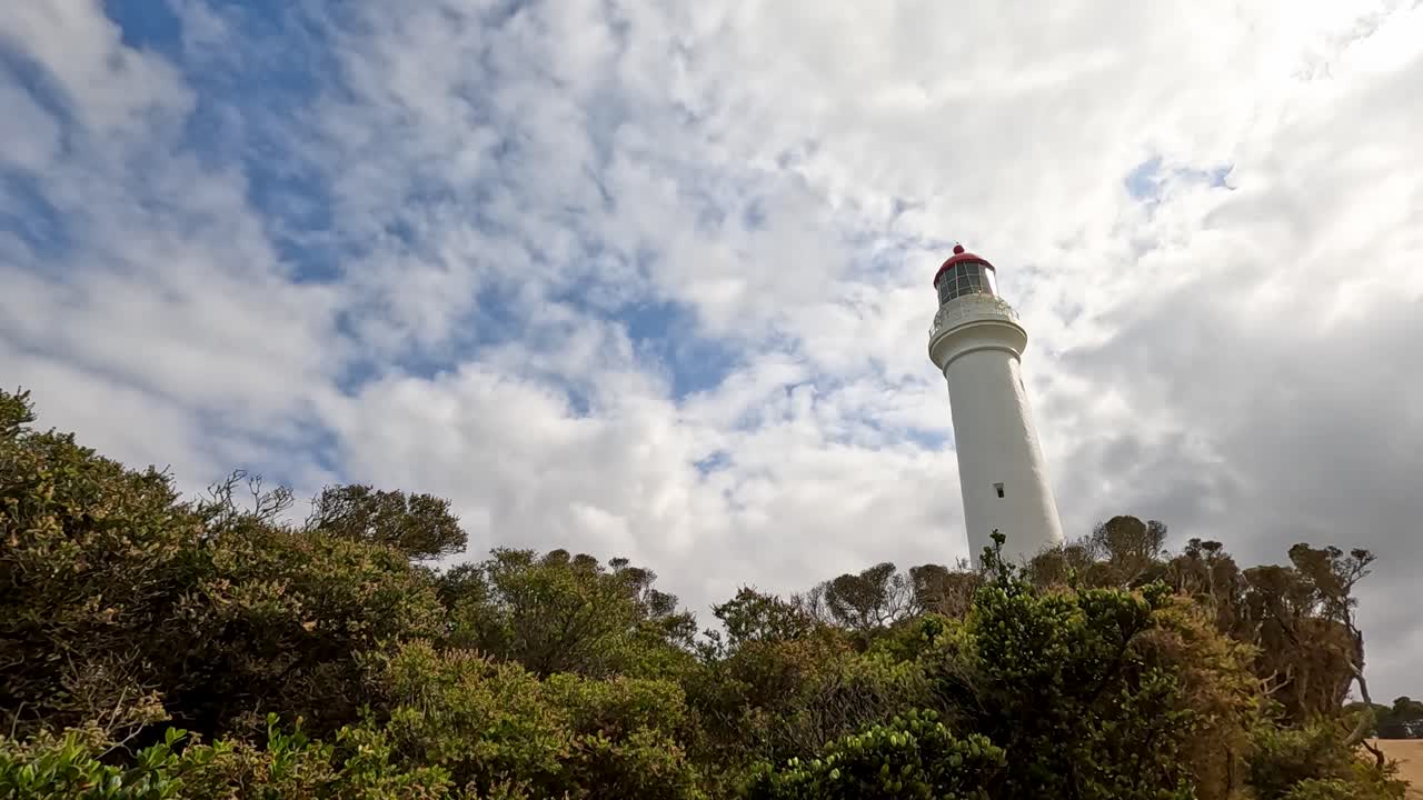 A lighthouse stands tall among dense greenery, captured under a dynamic, partly cloudy sky.