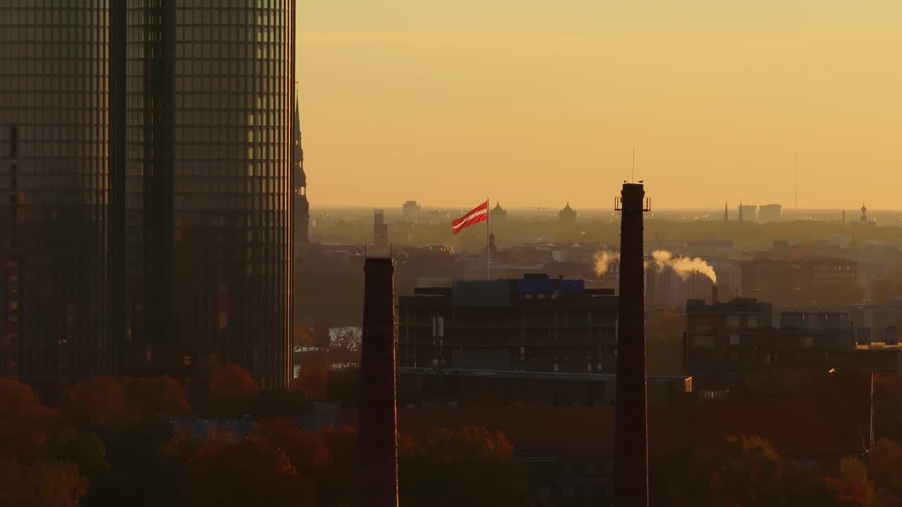 Drone glides left past Riga’s chimneys with waving Latvian flag and golden sky