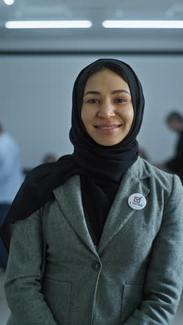 Una-Mujer-Se-Encuentra-En-Una-Mesa-Electoral-Moderna,-Posa,-Sonríe-Y-Mira-La-Cámara.-Retrato-De-Una-Mujer-Musulmana,-Votante-De-Las-Elecciones-De-Los-Estados-Unidos-De-América.-Fondo-Con-Cabinas-De-Votación.-Concepto-De-Deber-Cívico.