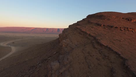 Drone-flying-over-rocky-formation-in-desert-with-red-mountain-in-background-at-sunset,-Morocco